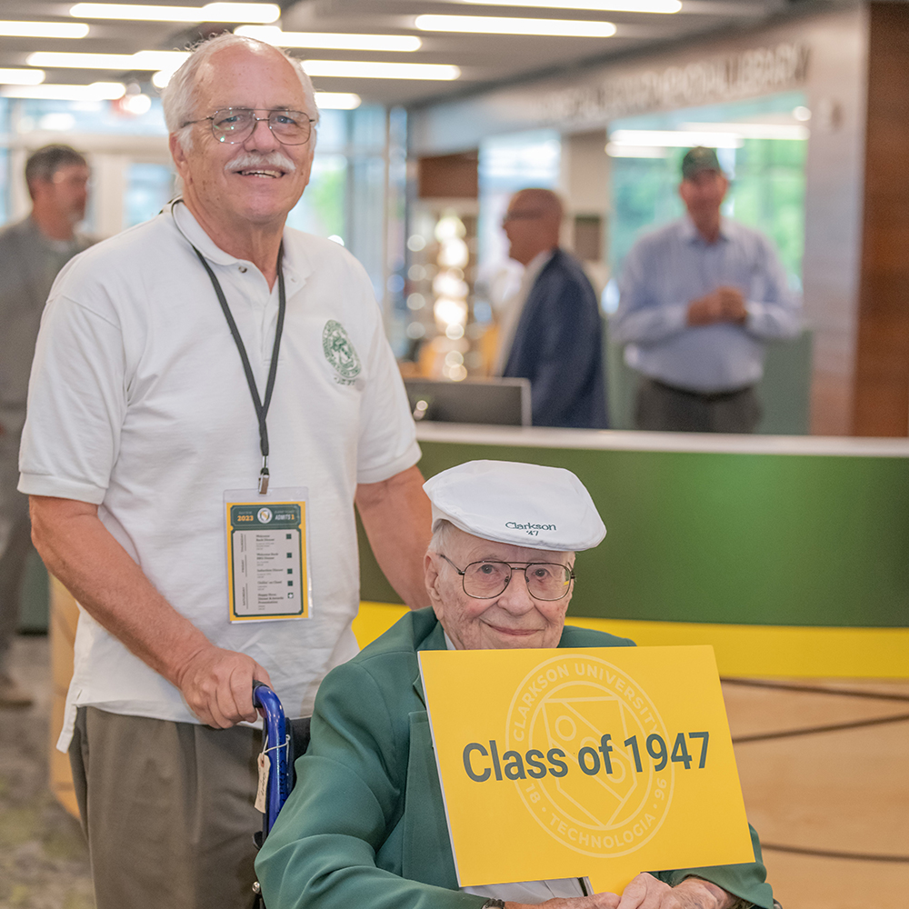 A man in a wheelchair holding a sign with Class of 1947 for the year he graduated. Another man is pushing the wheelchair.